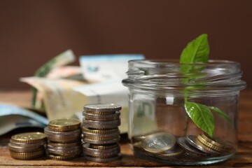 Glass jar with coins, sprout and banknotes on wooden table, closeup
