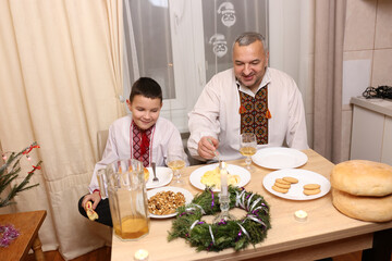 Father and son in embroidered shirts celebrate Christmas at a festive table with traditional food, bread and Christmas decorations.