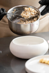 Chef plating risotto from stainless steel pot into modern white bowl. Culinary preparation in professional kitchen with black gloves and beige apron.