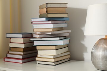 Many stacked books and lamp on white table indoors, closeup