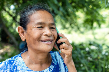 happy asian senior mature woman relaxing in garden, speaking on mobile phone with family, thai asia elderly grandmother enjoy smart outdoor lifestyle with wireless technology and digital device