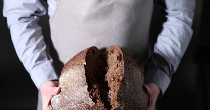 Baker breaking loaf of fresh bread on black background, closeup. Camera moving down