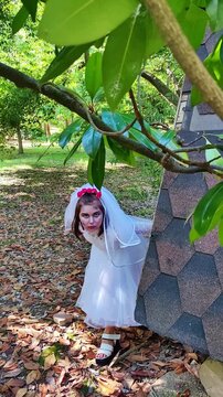 A girl in a white dress and a veil, the image of a scary bride, moves like a zombie on the street, in the park on Halloween