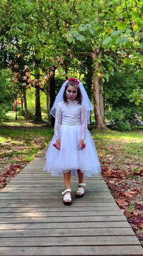 A girl in a white dress and a veil, the image of a scary bride, moves like a zombie on the street, in the park on Halloween