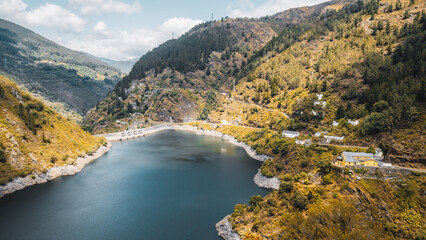 A serene mountain reservoir between steep forested slopes, concrete dam, and hillside houses with winding road under soft clouds, clear teal water, and rugged rocky shoreline