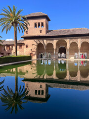 Andalusien, Alhambra in Granada mit maurischer Architektur, Palmen und Spiegelung im Wasserbecken unter blauem Himmel, faszinierende Reiseimpression aus S&uuml;dspanien