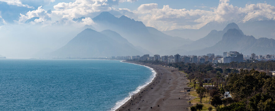 Panoramic view of Konyaaltı Beach's curved coastline with turquoise sea, white foam, and people relaxing against a backdrop of hazy Taurus Mountains. Sunny winter day in Antalya, Turkey.