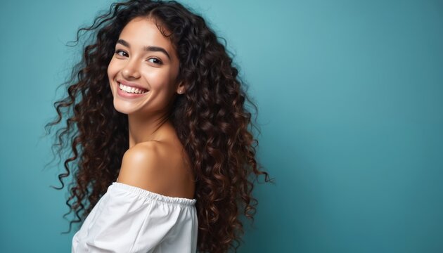 Attractive woman smiles. Female model looks over shoulder at camera in studio. Long curly hair is well groomed against a blue background. Beauty product advertising concept with copy space.