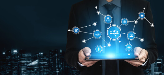 A businessman elegantly holds a tablet displaying various digital connections, symbolizing technology impact on modern business and communication in a night cityscape. Tessel