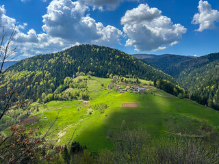 A scenic mountain village nestled in vibrant green hills and dense forest, under a dramatic sky with fluffy clouds casting shadows across the landscape.
