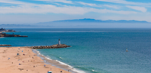 Santa Cruz beach features a sandy shore with visitors, a lighthouse on a rocky jetty, calm blue waters, a sailboat, and a hazy mountain backdrop.