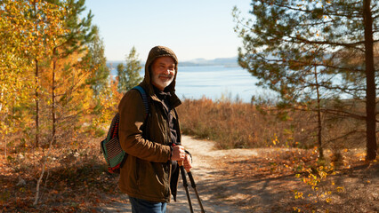 A senior citizen with trekking poles in his hands on a mountain slope route against the picturesque backdrop of autumn trees and a river below.