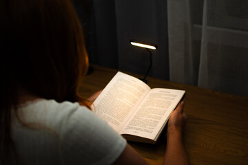 A woman is reading a book at a wooden table in the evening with the help of convenient clip-on lamp