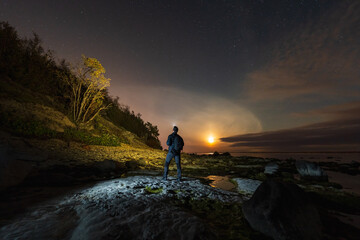 A lone hiker with a headlamp explores the rocky coastline of the Paldiski peninsula in Estonia under a starry night sky.
