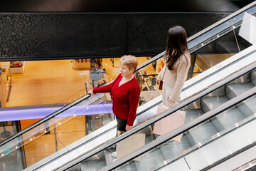 Two women walk down an escalator in a shopping mall.