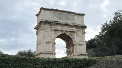 Fototapeta premium Rome, Italy - 13 January 2025. The Arch of Titus stands alone on a grassy rise, with its single barrel-vaulted passage, inscription, and carved reliefs still intact against a cloudy sky.