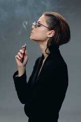Obraz premium Stylish woman smoking electronic cigarette with smoke on grey background, wearing black outfit and sunglasses