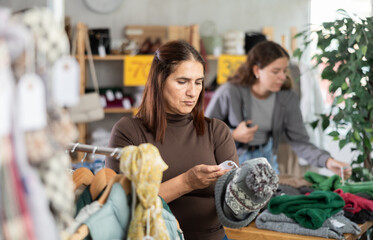 Latin woman customer examine and choose hat in store. Concept of seasonal wardrobe shopping. Client enjoys shopping for stylish clothes. Buyer browsing clothes goods in background
