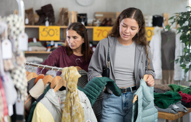 Smiling young female customer reviewing sleeveless winter jackets during shopping in retail outlet