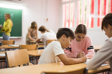 Teenage schoolchilds working in groups at lesson in secondary school with teacher on background