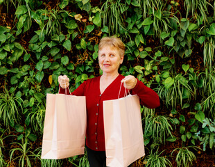 An elderly woman with bags in her hands stands near a wall with plants.