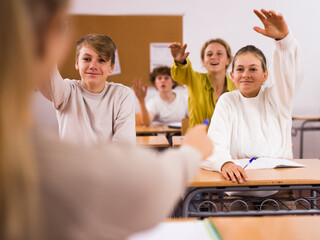 Teenage boys and girls sitting at desks in classroom and raising hands to answer question. Teacher pointing at girl.