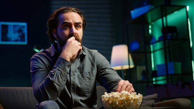 Puzzled man eating popcorn while watching confusing movie on streaming service, enjoying time off from work. Person consuming snacks, looking at film broadcasted on TV with convoluted plot, camera A