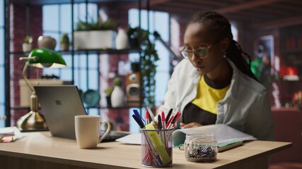 Bookkeeper in home office reviewing tax forms, reading reports on laptop and writing down information. Remotely working accountant managing finances remotely, taking notes, camera A