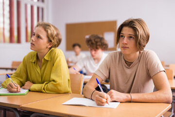 Fototapeta premium Teenage girl and boy sitting together at desk and doing tasks in classroom.