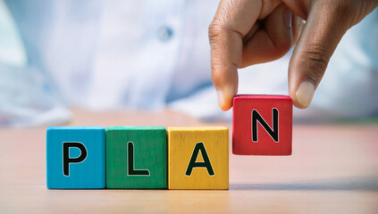 Close-up of colorful wooden blocks spelling PLAN with hand placing letter N, symbolizing strategy, organization, business planning, success, future vision, creativity, and achieving goals.