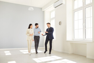 Fototapeta premium Professional realtor presenting new apartment to young family couple and discussing terms of purchase contract. Real estate agent showing clients new home or office space, talking about its benefits.