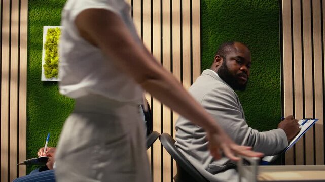 Confident job applicants filling in documents with their details, sitting in the hallway back to back before hiring interview. Reviewing CVs and preparing for employment questions with HR. Camera A.