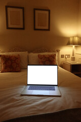 Laptop with blank white screen resting on a bed in a hotel room at night, showing work from home or relaxing concept. Mockup copyspace