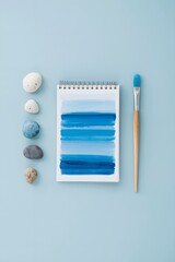 Top view of watercolor notebook with blue paint strokes, surrounded by brush and smooth stones on pastel blue background, symbolizing creativity, art inspiration, calm design concept, and minimalist w