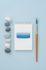 Top view of watercolor notebook with blue paint strokes, surrounded by brush and smooth stones on pastel blue background, symbolizing creativity, art inspiration, calm design concept, and minimalist w