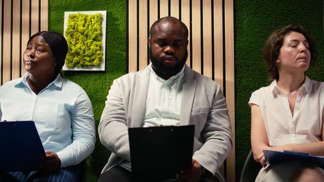 Professional diverse candidates awaiting start of job interview, showcasing anticipation in office hallway, preparing with information for the recruitment process. Career opportunity. Camera A.