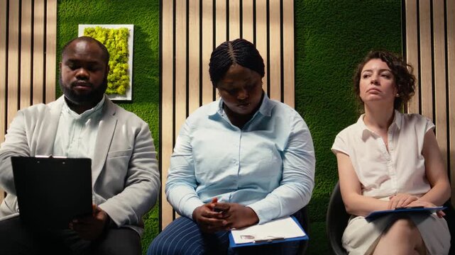Anxious diverse job seekers awaiting job interview discussion with manager, showcasing anticipation in office hallway. People preparing with information for the recruitment process. Camera A.