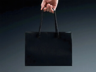 A hand holding a sleek black shopping bag with rope handles against a dark background.