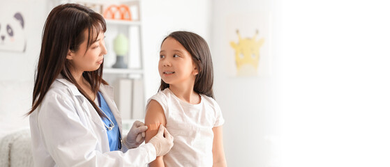 Fototapeta premium Female Asian doctor applying medical patch on little girl's arm after vaccination in clinic
