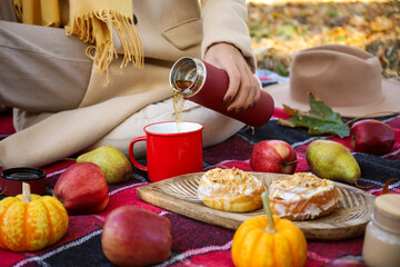 Young woman with fruits and buns pouring tea into cup on picnic in autumn park, closeup