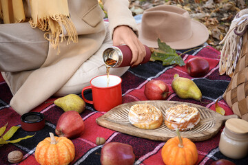 Young woman with fruits and buns pouring tea into cup on picnic in autumn park, closeup