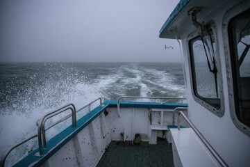 A boat navigates rough seas under a cloudy, grey sky, creating a frothy wake