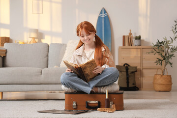 Cute teenage happy redhead girl with adventure book, world map and suitcase sitting at home