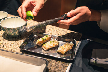 Final preparation of a traditional Japanese fish dish, with a chef grating fresh Japanese lime (sudachi or kabosu) over grilled fish fillets for added flavor. Authentic Japanese cuisine.