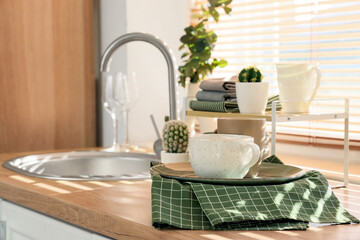 Folded clean towels with many dishes, cups and houseplants on counter near window in kitchen