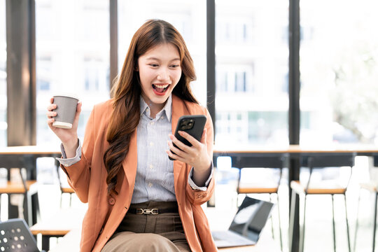 Excited young Asian businesswoman reacts happily while reading good news on her mobile phone and enjoying a coffee break in a modern office environment.