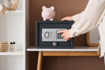 Woman with piggy bank opening modern safe box on table near brown wall, closeup