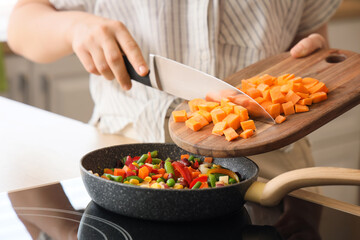 Woman putting cut carrot into frying pan in kitchen, closeup