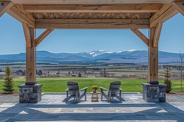 A scenic view from a wooden patio overlooking rolling fields and snow-capped mountains, featuring two chairs and a small table with flowers.