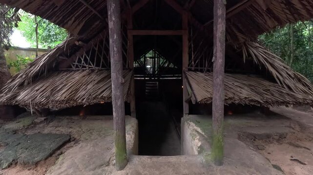 Entrance to an underground room at the Cu Chi Tunnels in Vietnam, with a traditional thatched roof shelter and narrow earthen pathway.